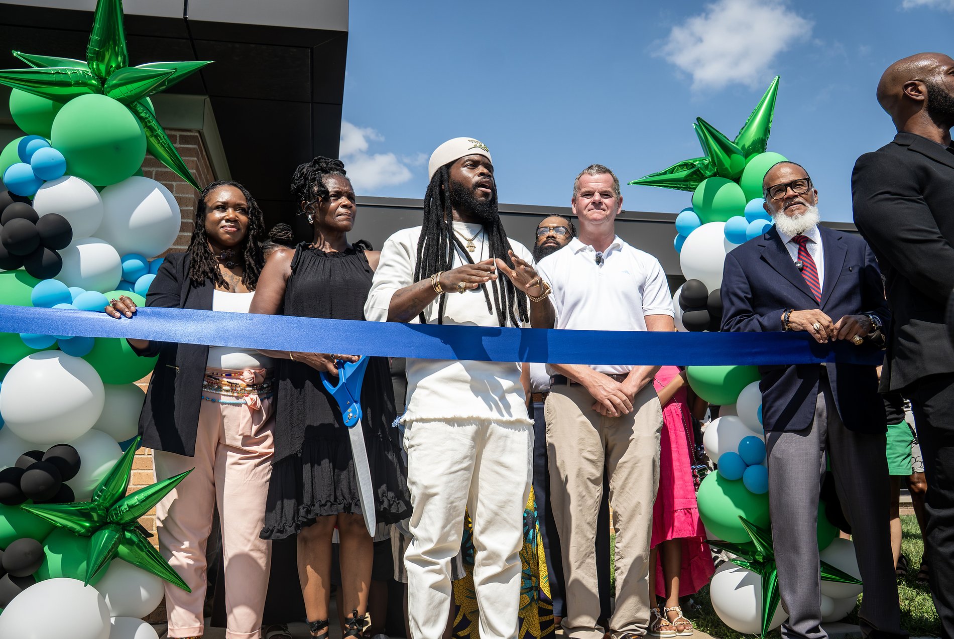 Dr. Yah'ki speaking at the ribbon cutting for the Center, standing at the middle of a line of family, staff and civic guests behind a blue ribbon, with green, blue, white and black balloon arches framing both sides of the entrance.